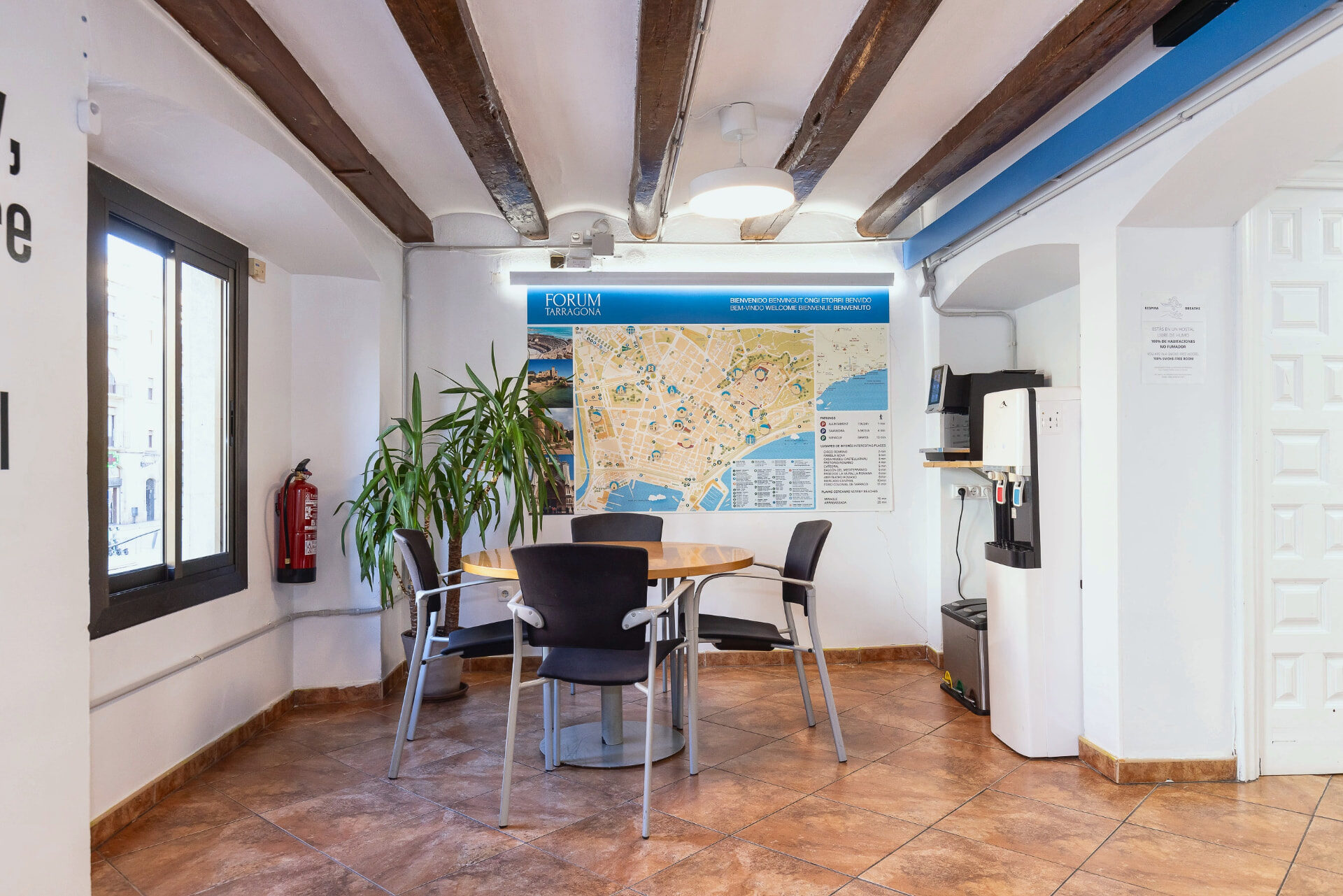 Taula amb cadires i mapa de Tarragona a la sala comuna, al costat de les finestres a la plaça.